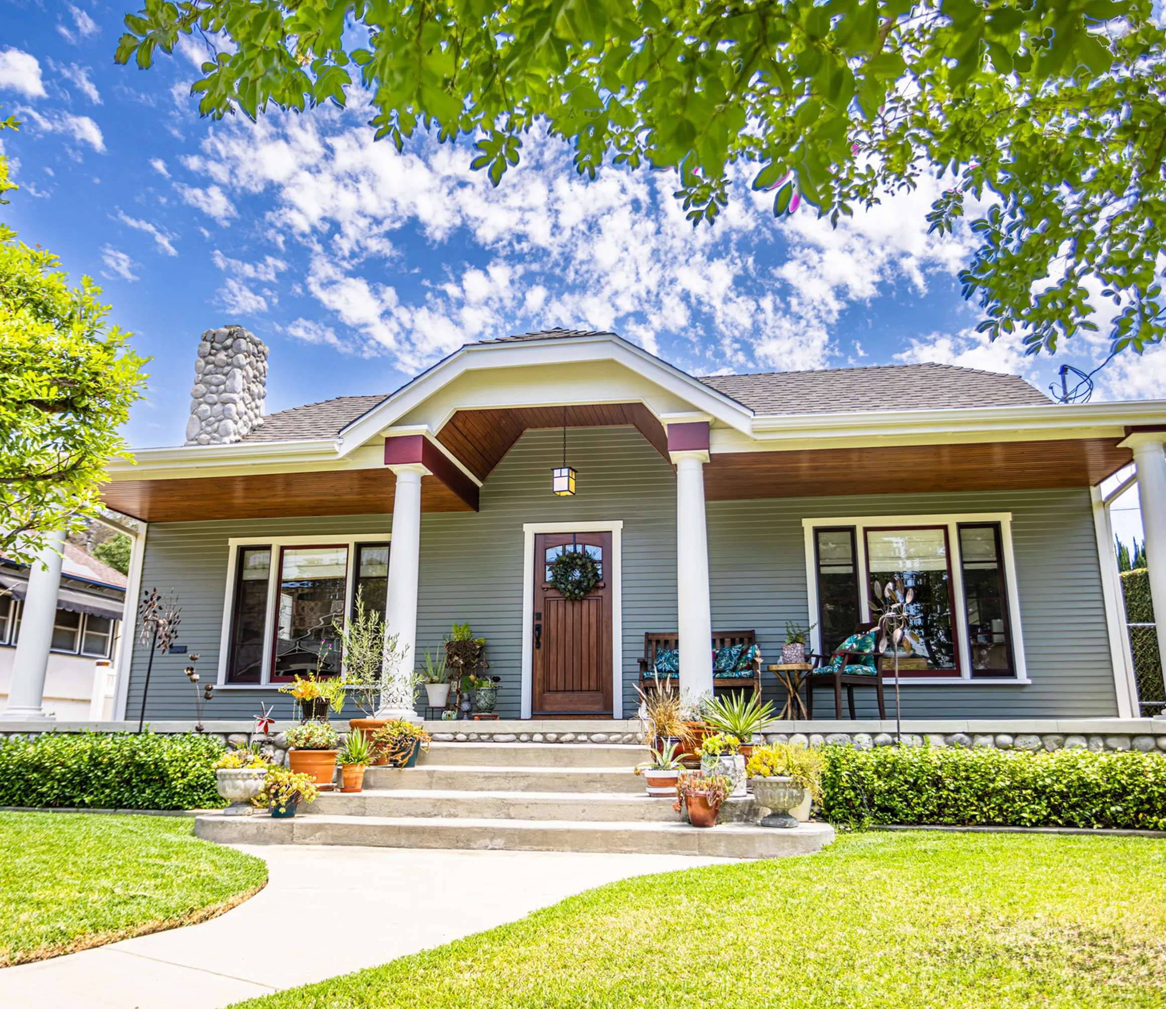 Bungalow-style house with a front porch, wooden door, and stone chimney, surrounded by a manicured lawn and plants. Bright blue sky with scattered clouds overhead.