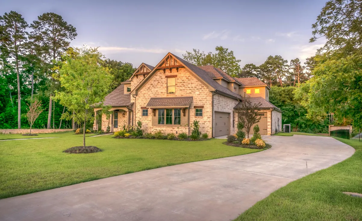 A large brick house with a well-maintained lawn, surrounded by trees. A long driveway leads to a two-car garage.