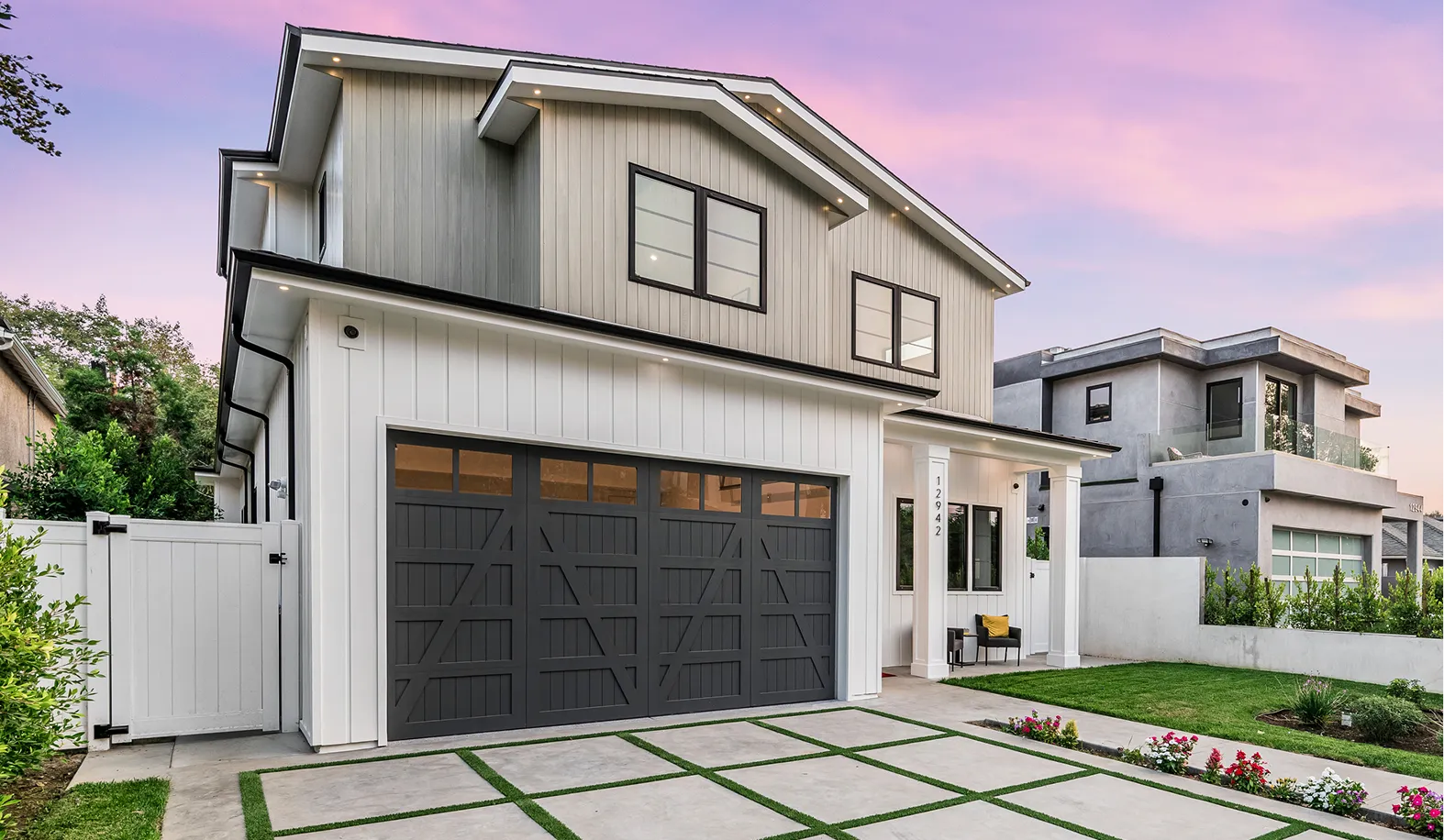 Modern two-story house with gray accents, a dark garage door, and a small front yard. The sky is pinkish at sunset.
