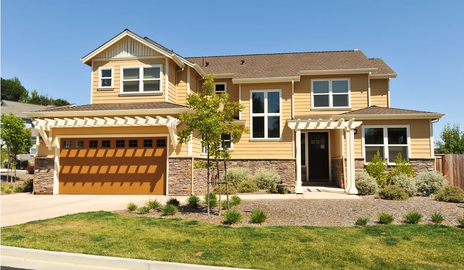 A two-story beige house with a wooden garage door, stone accents, and a small front yard with green grass and shrubs, under a clear blue sky.
