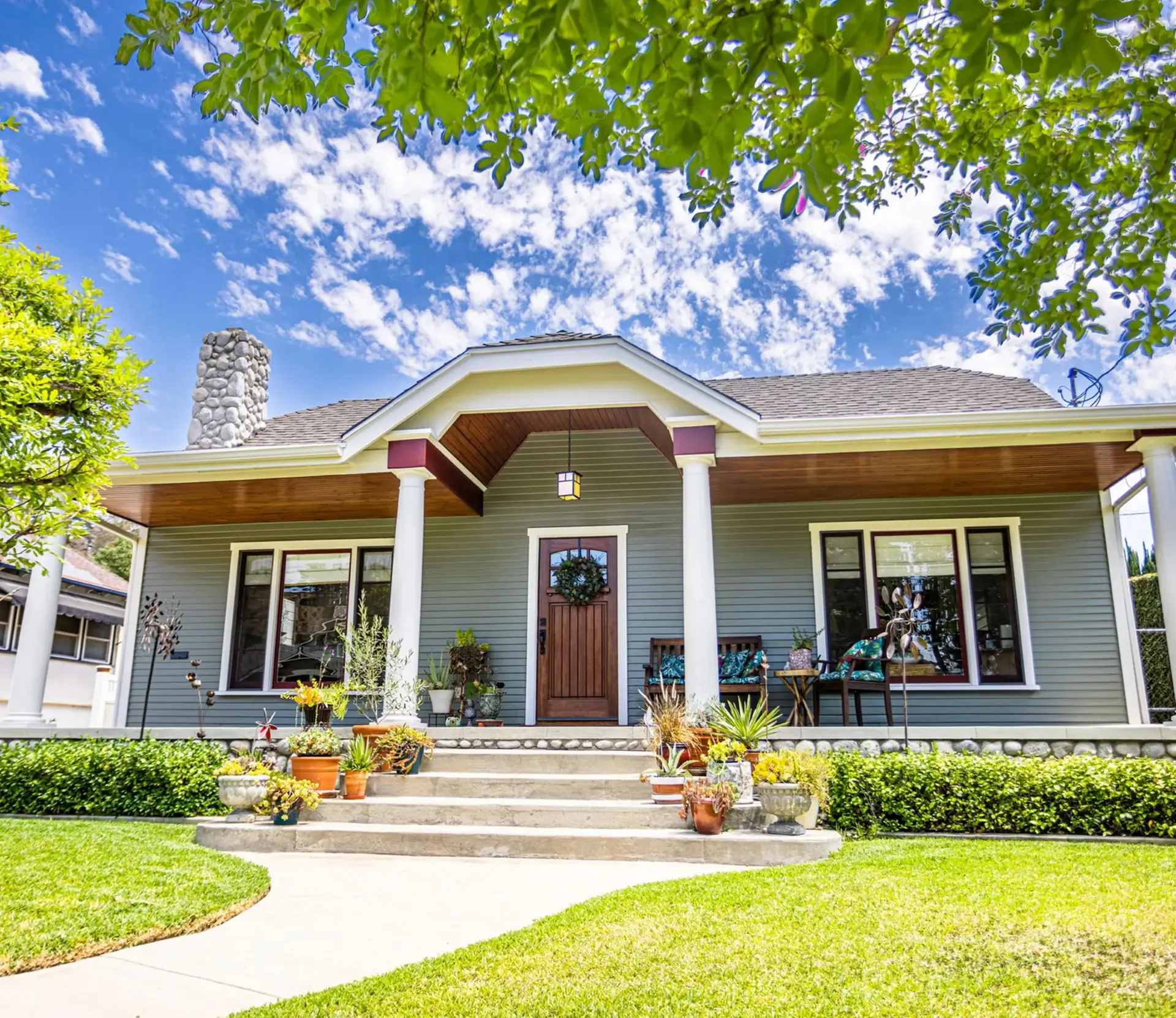 Bungalow-style house with a front porch, wooden door, and stone chimney, surrounded by a manicured lawn and plants. Bright blue sky with scattered clouds overhead.
